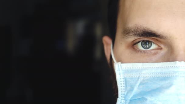 Porträt eines jungen Mannes mit medizinischer Maske blickt während der Coronavirus-Pandemie mit müden Augen in die Kamera. — Stockvideo
