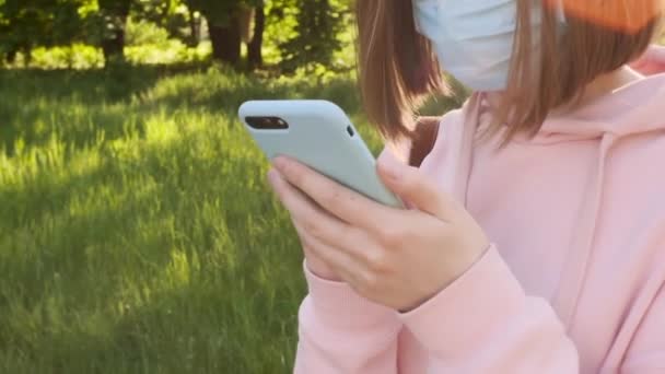 Una joven con una máscara médica protectora utiliza un mensaje de texto para teléfonos inteligentes, pandemia de noticias de búsqueda, autoaislamiento . — Vídeo de stock
