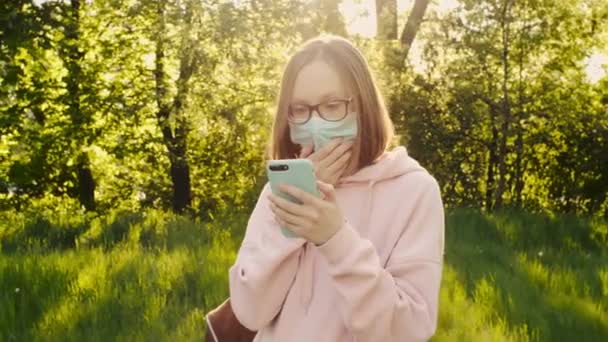 Choqué jeune fille dans un masque médical de protection regarde l'écran d'un smartphone . — Video