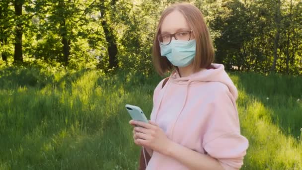 Portret młodej modnej dziewczyny w medycznej masce ochronnej ze smartfonem w parku. — Wideo stockowe