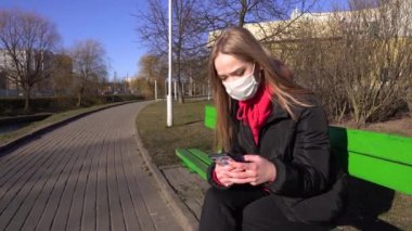 Koruyucu maskeli bir kız bankta oturuyor ve şehir parkında telefonla konuşuyor. Salgın Covid-19 koronavirüs koruması. Nehir kenarındaki bir bankta dinlenmek için oturdum. 4k
