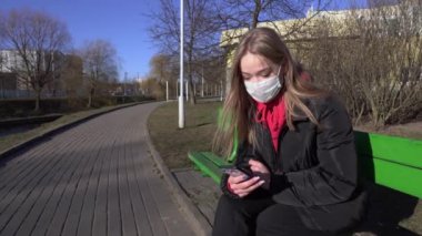 Koruyucu maskeli bir kız bankta oturuyor ve şehir parkında telefonla konuşuyor. Salgın Covid-19 koronavirüs koruması. Nehir kenarındaki bir bankta dinlenmek için oturdum. 4k