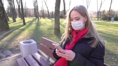 Koruyucu maskeli, kırmızı elbiseli genç bir kadın şehir parkında video kaydetmek için akıllı telefon kullanıyor. Salgın Covid-19 koronavirüs koruması. Nehir kenarındaki bir bankta dinlenmek için oturdum. 4k
