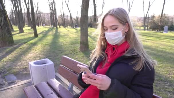 市内の公園でビデオを呼び出すか 記録するためにスマートフォンを使用して保護マスクの赤で若い女性 パンデミックCovid 19コロナウイルス保護 私は川のそばのベンチに腰を下ろした — ストック動画