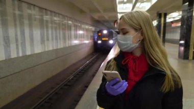 Genç sarışın bir kız metroyu bekliyor ve telefonu kullanıyor, bulaşıcı bulaşıcı hastalıklara karşı koruyucu bir maske takıyor ve toplu taşımacılıkta gribe karşı bir koruma olarak. Coronavirus 4k
