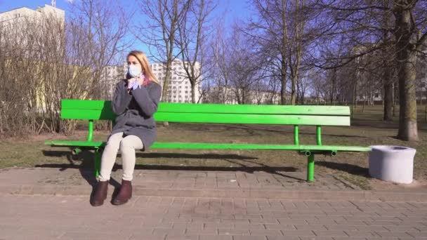 若い女性は マスクと手袋の上に置くベンチに座っている日没時にコロナウイルスCovid 19の検疫パンデミックの際に都市公園内の電話を使用4K — ストック動画