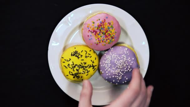 Αρσενικό χέρι παίρνει από ένα λευκό πιάτο χρώματος κίτρινο, ροζ και μοβ ντόνατς σε μαύρο φόντο. Άνω όψη — Αρχείο Βίντεο