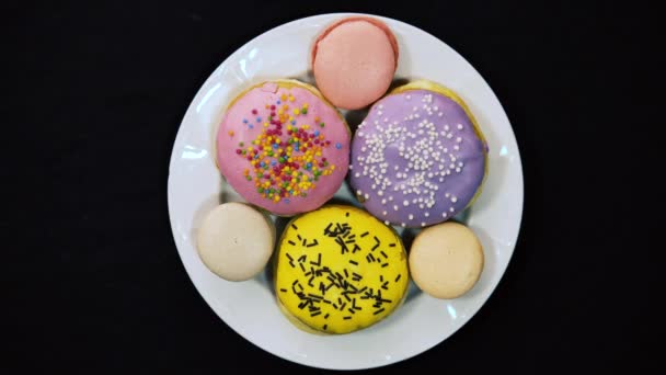 Lapso de tempo em uma placa branca deliciosos donuts de chocolate multi-coloridos e macaroons coloridos em um fundo preto 4k Gingerbreads na forma de um donuts mentira em uma placa de vidro branco — Vídeo de Stock