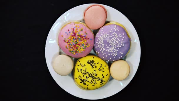 Időeltolódás egy fehér tányéron finom csokoládé sokszínű fánk és színes macaroons fekete alapon 4k mézeskalács formájában fánk fekszik egy fehér üveglapon — Stock videók