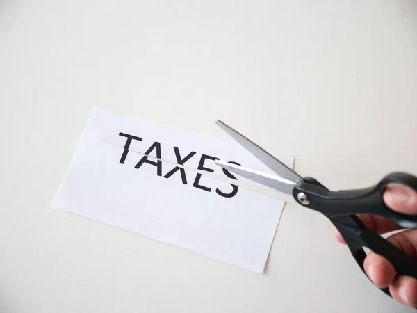 Closeup photograph of man holding black handle scissors and cutting a paper with the word taxes on it representative of federal and state tax cut reform saving people money with new deduction policy.