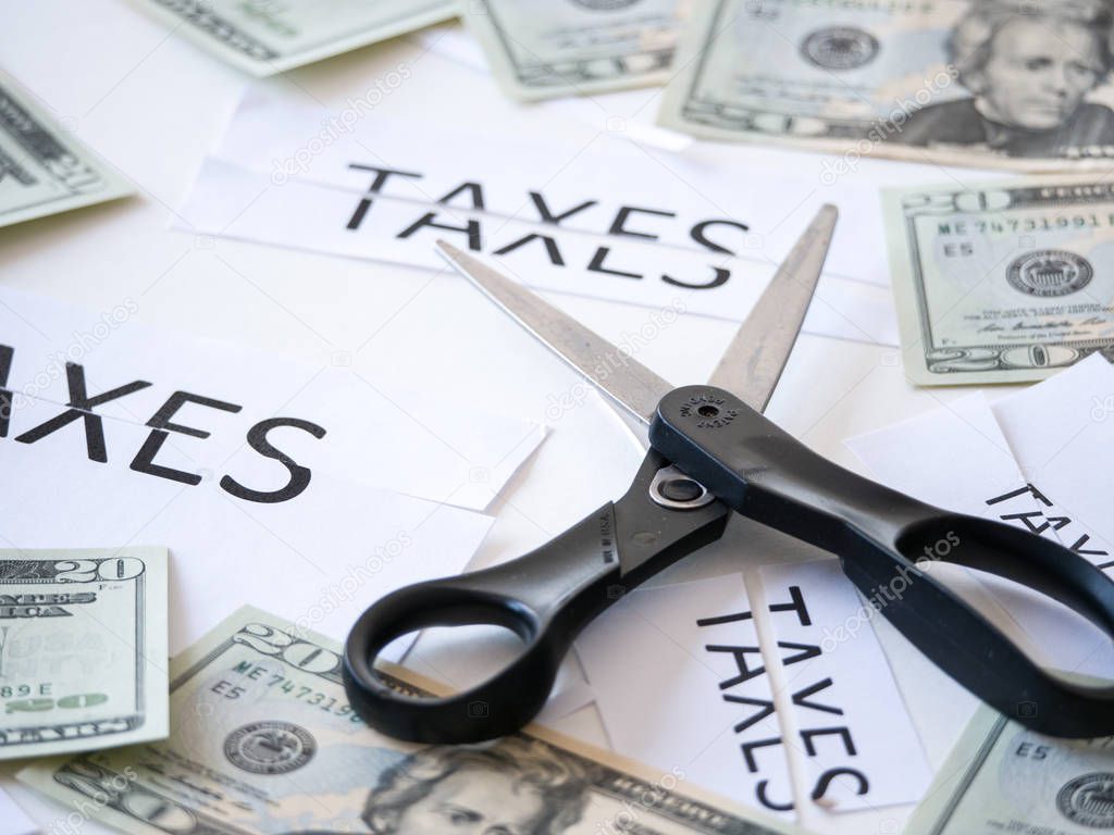 Close up photograph of a scissors cutting a paper that has taxes typed on it with 20 dollar US money representative of government tax cuts and saving money.