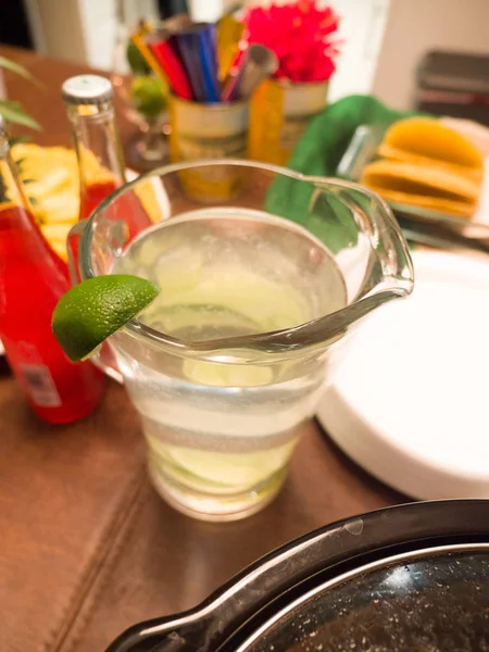 Close up of a full glass water pitcher with lime juice and green lime slice on top of pitcher on a dining table with Mexican themed party decor in background.