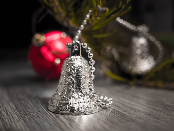 A closeup photograph of silver Christmas bells on a beaded strand on a gray woodgrained floor and pine needle branch and red ball ornament in background making a festive holiday background.