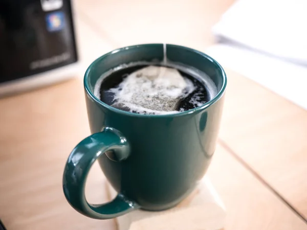 Close up photograph of a hot coffee mug full of water and a tea bag on top of a coaster and wood desk at an office work station with computer and papers in the background.