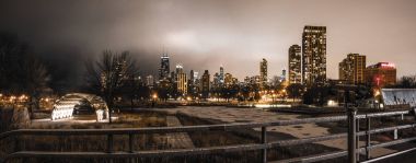 Chicago, IL - January 23, 2018: Panoramic view of the Chicago skyline at night from the bridge overlooking Lincoln Park's South Pond near the zoo on a still winter night with snow on the ground and lights on buildings and gray sky clipart