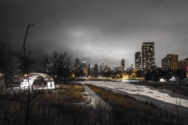 Panoramic landscape view of the Chicago city skyline from the bridge near Lincoln Park's South Pond adjacent to the zoo at night with snow covered paths below and partially frozen pond in foreground. clipart