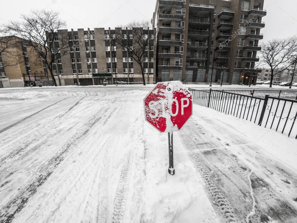 View of snow covered red stop sign with buildings, slippery shoveled concrete side walk and plowed snowy street beyond in Chicago during winter weather snow fall.