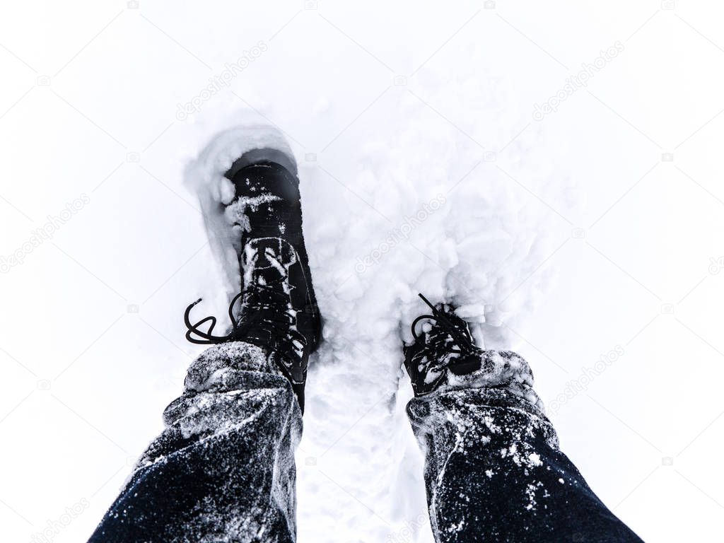 Close up view looking down at a pair of man's black colored insulated winter boots and snow covered blue jeans as he walks through the deep snow drifts in Chicago after snow storm weather in winter.