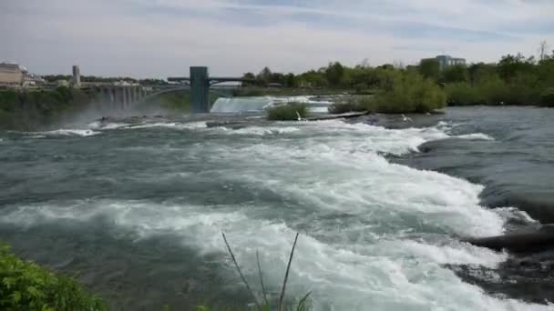 Weergave van haasten water rapids op Niagara rivier alvorens over het rotsmassief van American Falls waterval met blauwe lucht en witte wolken boven en weergave van de observatie dek en Canada aan de horizon. — Stockvideo