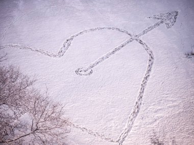 Photograph high above a large heart shaped temporary nature art installation with an arrow through it drawn in the fresh snow by a person stamping down the snow with grass showing through. clipart