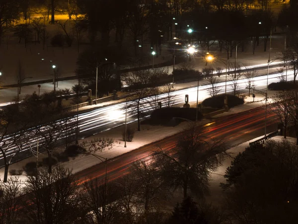 Long exposure photograph of car headlight and tail light streaks or trails on Lake Shore Drive in Chicago over foster beach underpass with speed limit sign and bare trees in snow covered ground.
