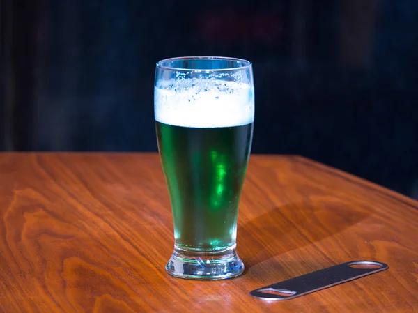 A pint glass of green beer with foamy white head celebrating the St. Patrick's day holiday sitting on a dark wood grain table and dark black backdrop.