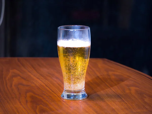 Photograph of a pint of light lager beer on a black walnut wood grain table top with dark background.