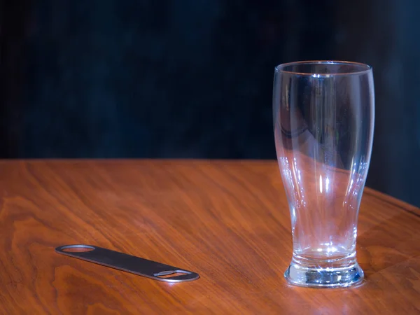 Photograph of an empty pint beer glass and stainless steel professional bottle opener bar tool on a black walnut wood grain table top with dark background.