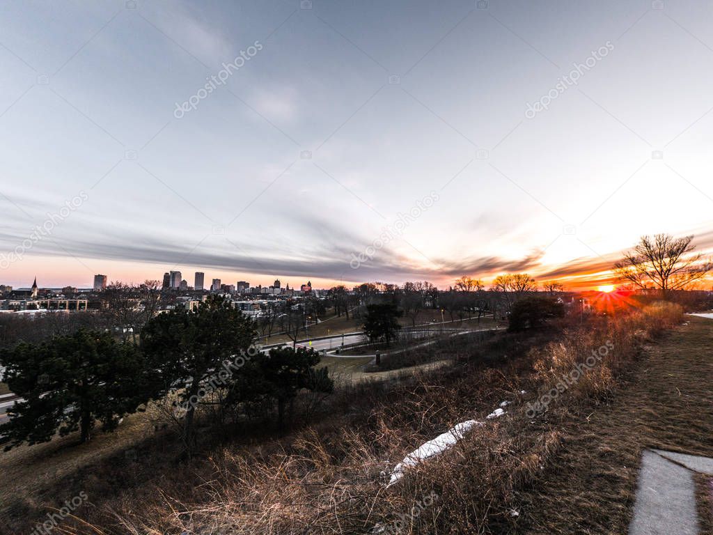 A beautiful landscape photograph of downtown Milwaukee at sunset with clouds in the sky and buildings and Lake Michigan at the horizon and trees and grasses in foreground at Kilbourn Reservoir Park.