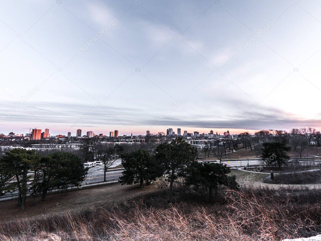 A beautiful landscape photograph of downtown Milwaukee at sunset with clouds in the sky and buildings and Lake Michigan at the horizon and trees and grasses in foreground at Kilbourn Reservoir Park.