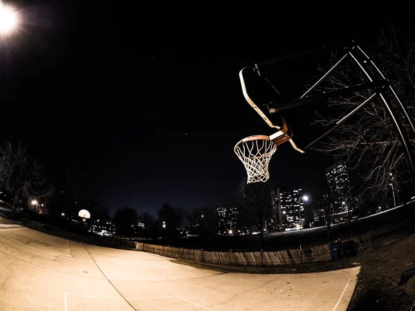 Night photograph of an outdoor weathered basketball court and basketball hoops with white backboards orange rims and nets lit with nearby street light in the city of Chicago.