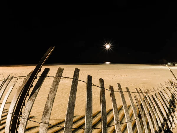 Night photograph of foster beach with weathered wooden lat and wire snow fence tipped over on its side in the sand with black dark Lake Michigan and the moon reflecting on water in the sky beyond.