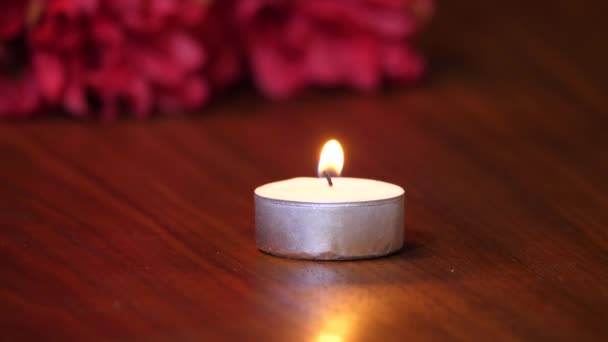 Vídeo de fondo de ambiente romántico de una vela de té con llama parpadeante en una mesa de grano de madera oscura y flores rojas en el fondo . — Vídeos de Stock