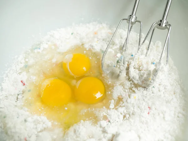 Close up photograph of Eggs and cake batter in a white ceramic mixing bowl with metal beaters, flower, oil, sugar and sprinkles in the batter.