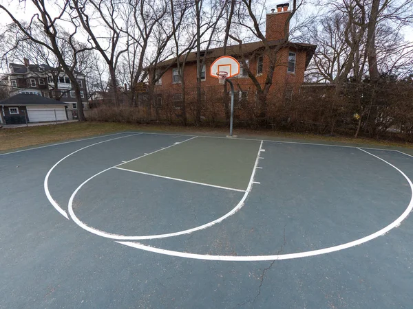 Photograph of an urban or city basketball court from the three point line arch looking towards the white and orange basketball backboard and hoop mounted to a metal post with buildings beyond.