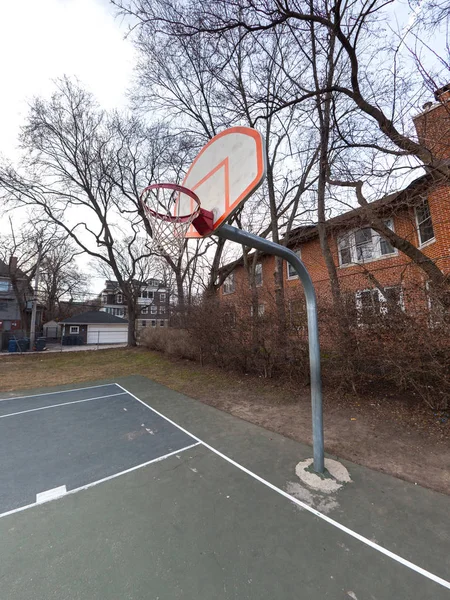 Photograph of an urban outdoor basketball court and white and orange backboard, rim and net mounted to a metal post with blue and green painted court and buildings and trees beyond.