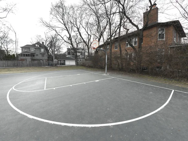 Photograph of an urban or city basketball court from the three point line arch looking towards the white and orange basketball backboard and hoop mounted to a metal post with buildings beyond.