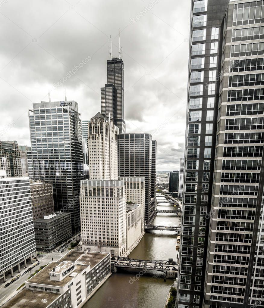 View of downtown Chicago from a high rise looking towards Willis Tower formerly Sears Tower with the dark green Chicago River and bridges below and cloudy sky above.