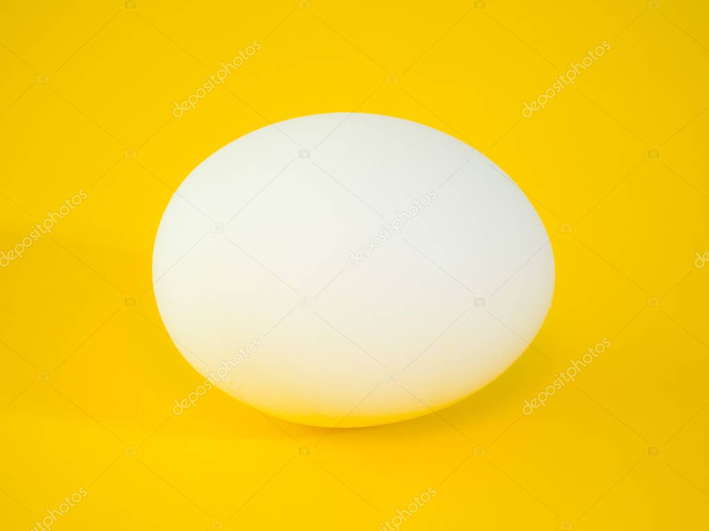 Close-up photograph of a real large white chicken egg on vibrant yellow solid color cutting board background making a beautiful eye catching background for the easter holiday.