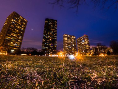 Night photograph of highrise condo buildings in Edgewater neighborhood in Chicago with purple and blue hue to the sky just after sunset with street and building lights on and grass in the foreground. clipart