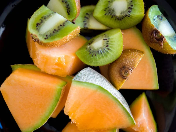 Close up food photograph of fresh ripe and mouth watering cantaloupe or muskmelon pieces with sliced kiwifruit served in a black bowl with vibrant orange and green colors.