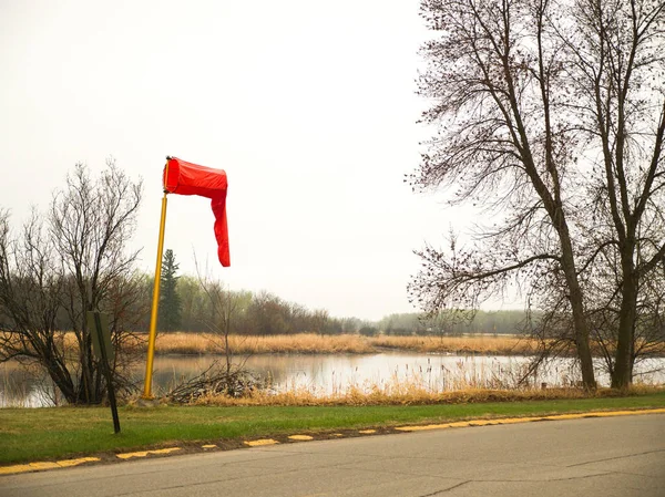 A bright red fabric wind sock on top of a yellow pole hangs limp along the river shoreline on a calm morning in Warroad Minnesota with trees and cloudy white and gray sky beyond.