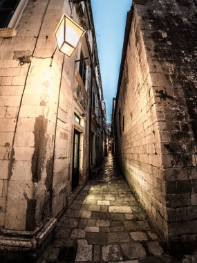 Beautiful night street photograph looking down a narrow stone paved pedestrian alley in the historic town of Dubrovnik in Croatia between buildings. clipart
