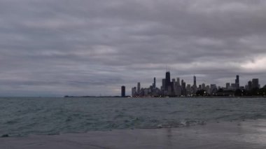 Dalgalar takviyeli beton kıyı şeridine yuvarlanıyor, yukarıda bulutlu bir gökyüzü ve ilerideki Chicago silueti gece çökerken ve dışarısı kararırken..