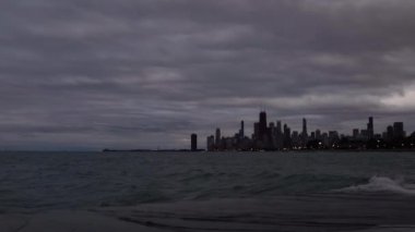 Dalgalar takviyeli beton kıyı şeridine yuvarlanıyor, yukarıda bulutlu bir gökyüzü ve ilerideki Chicago silueti gece çökerken ve dışarısı kararırken..