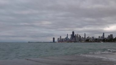 Dalgalar takviyeli beton kıyı şeridine yuvarlanıyor, yukarıda bulutlu bir gökyüzü ve ilerideki Chicago silueti gece çökerken ve dışarısı kararırken..