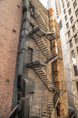 A view looking up a black painted steel fire escape adjacent to weathered and rusty ductwork on a multi story brick building in an urban environment surrounded by highrise buildings in Chicago. clipart