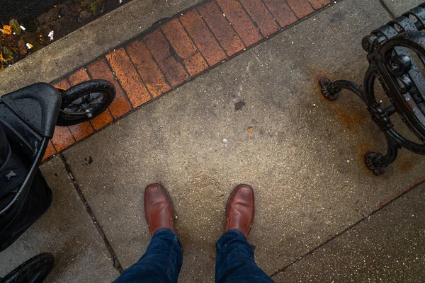 Looking down at the feet of a man wearing jeans and brown leather shoes standing on the concrete and brick paver sidewalk next to a baby stroller and a metal park bench.