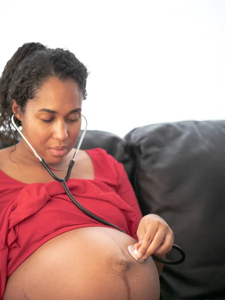 A beautiful young African American mixed race woman with dark curly hair in a pony tail and red dress listens to her unborn baby\'s heart beat using a stethoscope on her bare skin belly.