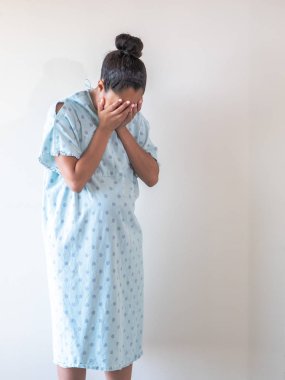 A young pregnant mixed race African American woman wearing a hospital gown or robe puts or hides her face in her open palms as she hunches over crying tears of sadness or depression. clipart